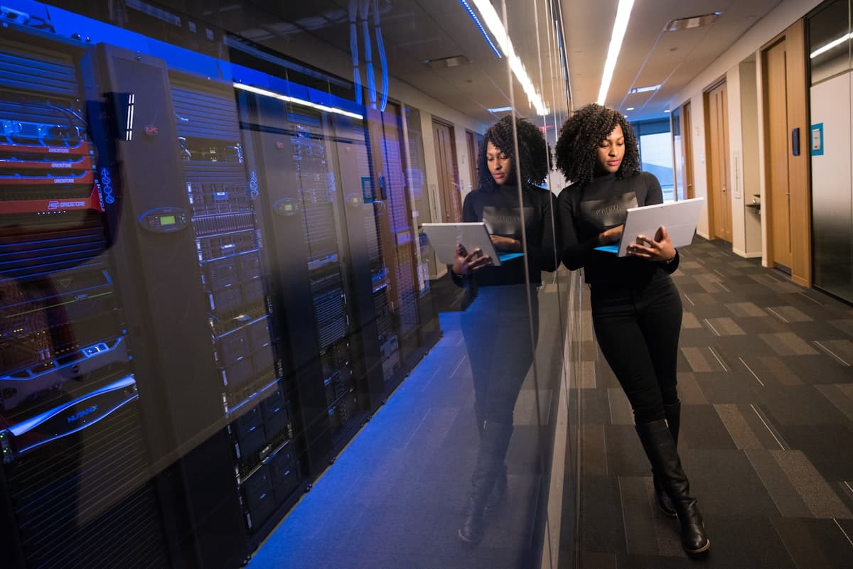 Woman holding a laptop next to a glass-enclosed server rack in a data center corridor