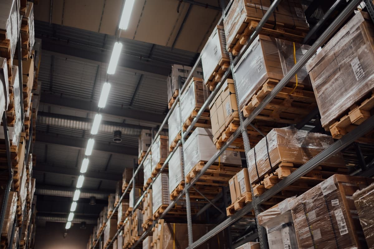 Tall warehouse shelving stacked with palletized cardboard boxes under fluorescent lights